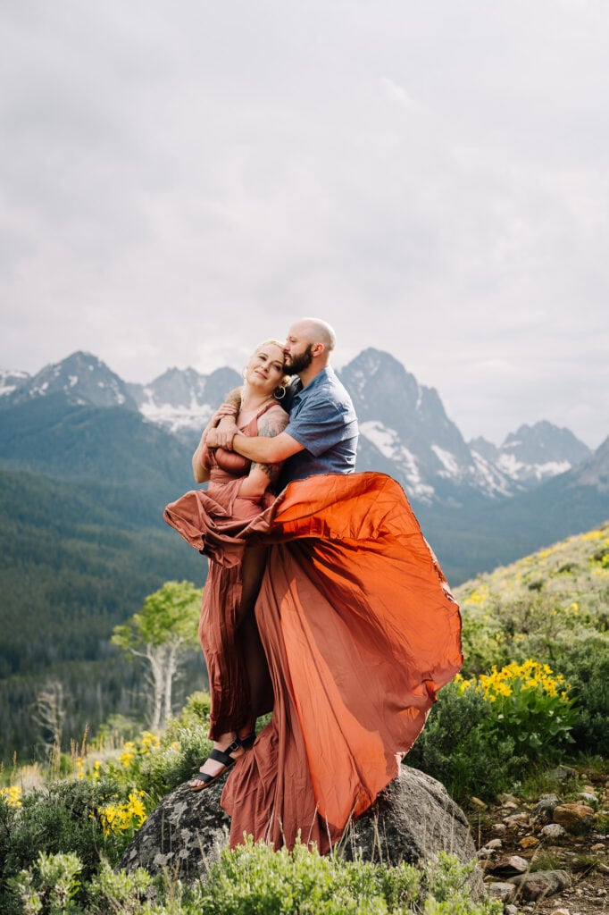 A couple stands together on a rock. There are mountains behind them. The man is hugging his fiance and her dress is blowing in the wind. This couple hiked to this location for their engagement session in the Stanley, Idaho mountains.