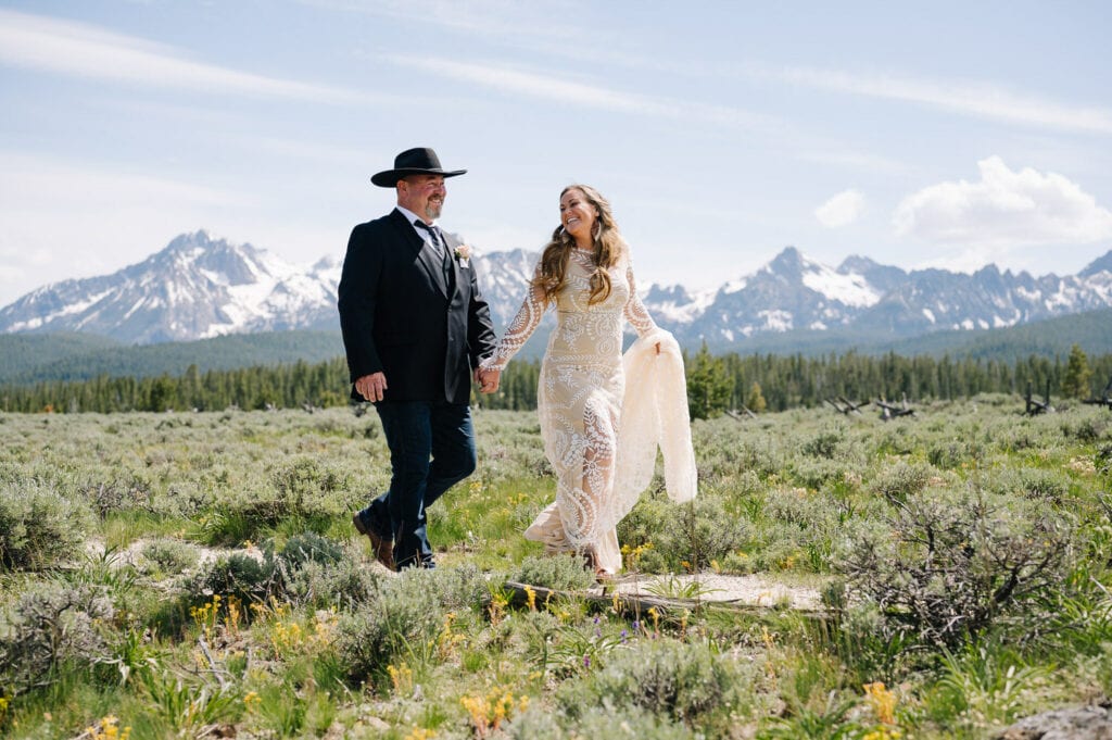 An eloping couple walks down a path after having their first look. There are mountains beyond the couple. The groom is wearing a black suit jacket and cowboy hat. The bride is wearing a long sleeve Rue De Shine style dress.