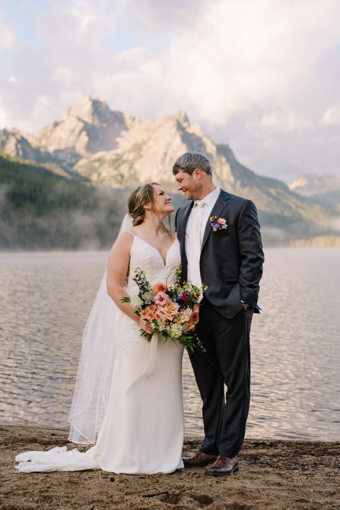 An eloping couple stands on the shore of an alpine lake in Idaho. There are mountains covered in a light fog behind them. The groom is wearing a dark grey suit and the bride is wearing a white dress with a long veil. 