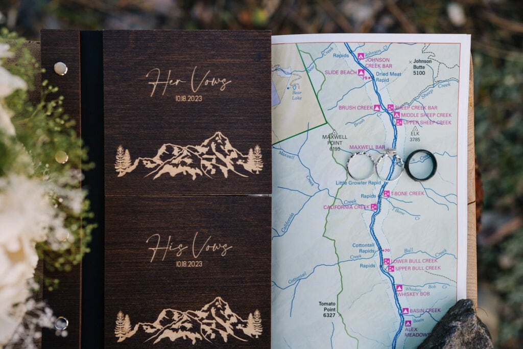 Dark wooden vow books lay face up next to a map. The vow books have 'his vows' and 'her vows' printed on the front. There is a map of the Salmon River in Idaho laying next to the vow books. There are wedding bands laying on the map.