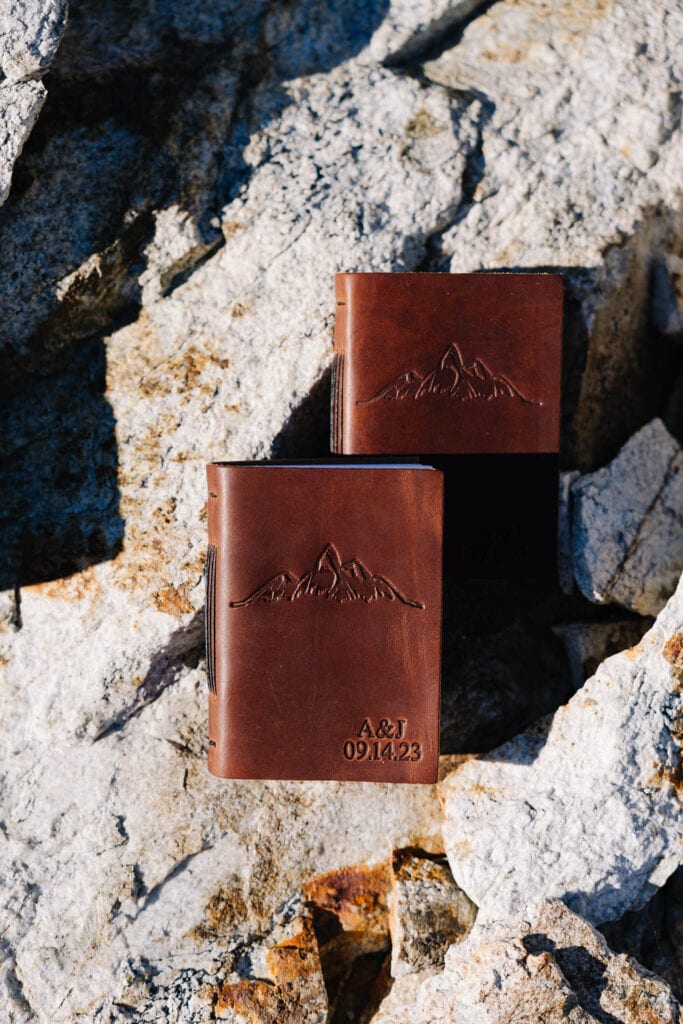 Two brown leather vow books lay on the ground in the sunshine. The vow books are customized with mountains, the couples wedding date, and the couples initials.