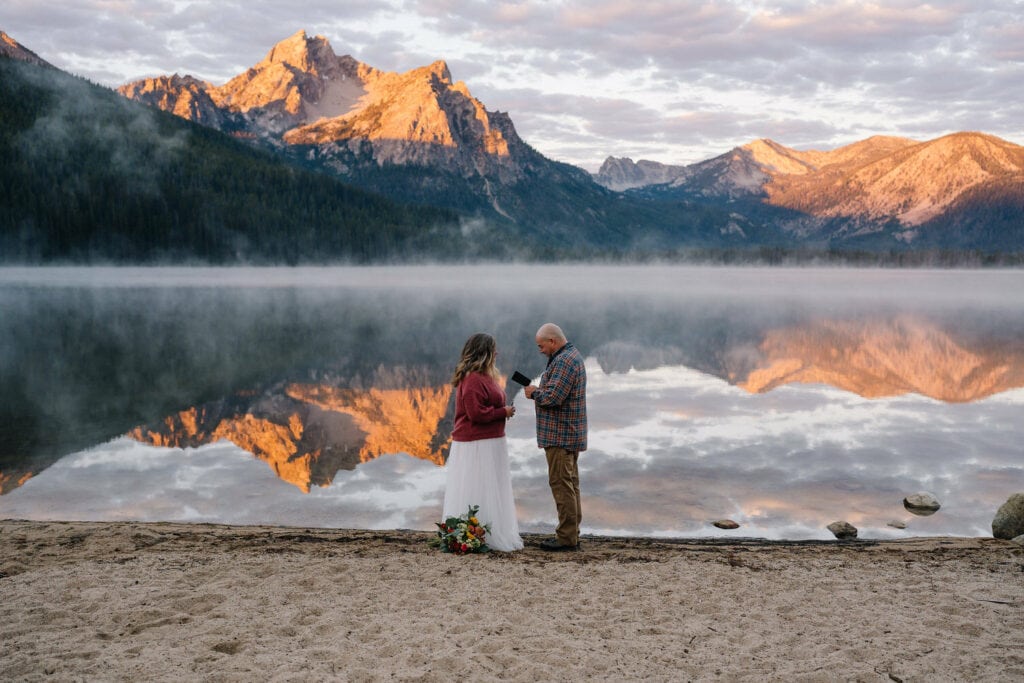 A groom reads vows to his bride at sunrise. The couple is standing along an alpine lake in Idaho. The bride is wearing a pink shirt with a white tule shirt.