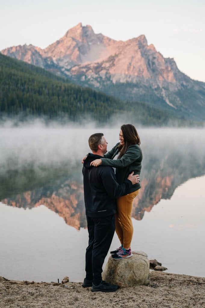 A couple stands next to a mountain lake. There is an alpenglow on the mountains behind the lake and mist is rising off of the water. This couple chose a sunrise location for their engagement session in Stanley, Idaho.