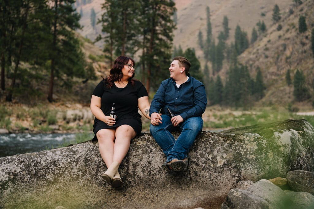 A couple sits on a rock along the Salmon River. They are holding hands and smiling at each other.