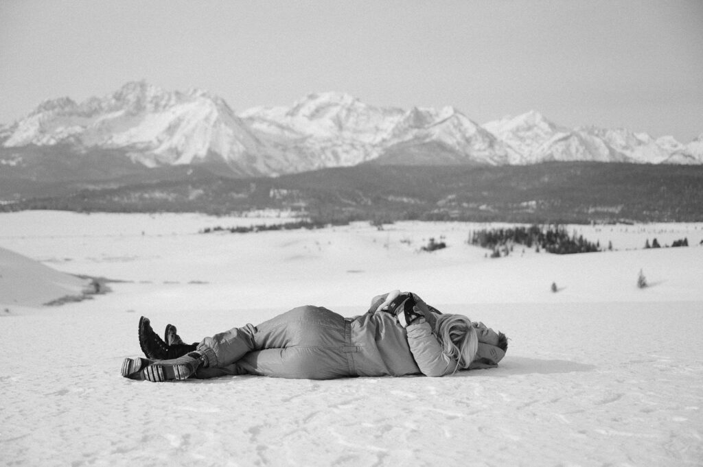 A couple lays on the snowy ground together during their engagement session. Their faces are away from the camera and they are looking at a frozen mountain landscape.