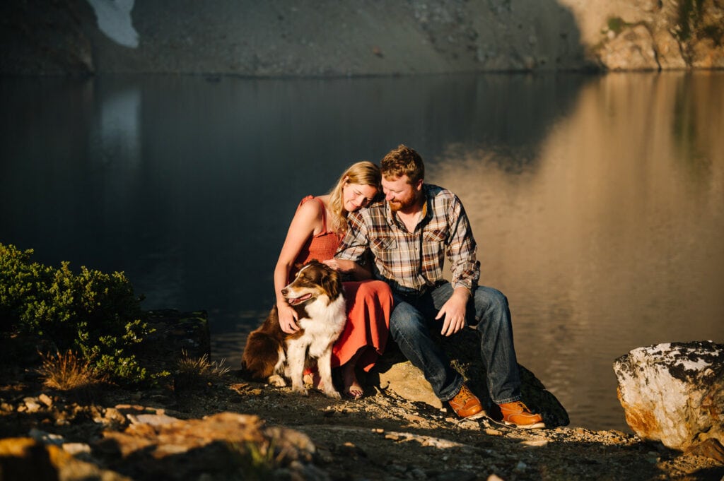 A couple sits with their dog in the early morning sun. The couple is wearing blue and orange tones. This couple hiked to a lake for their sunrise engagement session in Boise.