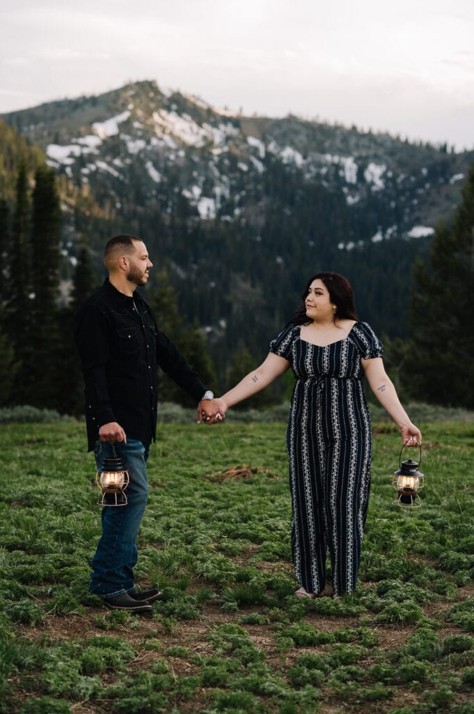 A couple walks holding hands in a mountain meadow at dusk. The couple is holding lanterns in their outside hands. This couple wore black and grey toned clothing for their engagement session in Boise.