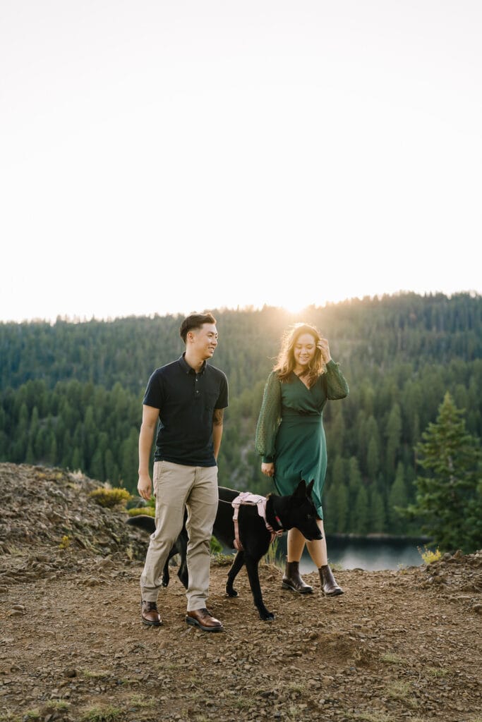 A couple walks with their dog during their engagement session at Ponderosa State Park in McCall. The couple is wearing blue and green toned clothing. The sun is peaking over the mountains and highlighting their silhouettes.