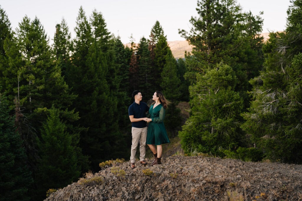 A couple stands together on a rock outcropping during their engagement session in Ponderosa State Park. There are pine trees behind them. This couple wore green and blue toned engagement oufits.