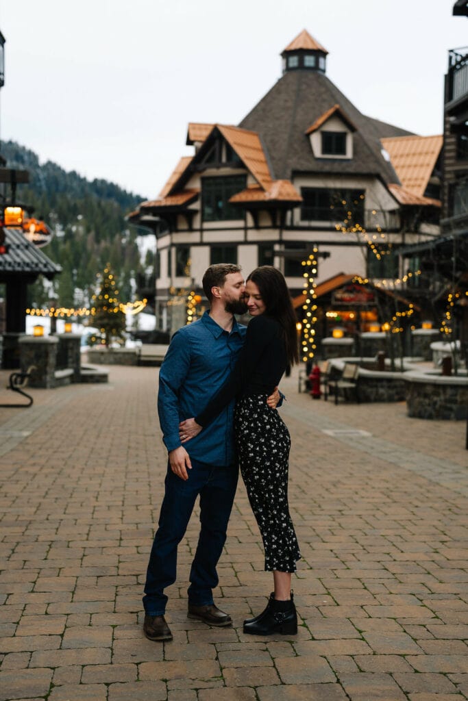 A couple stands in the Village at Tamarack Lodge during their engagement session in McCall. He is kissing her cheek and she is smiling.