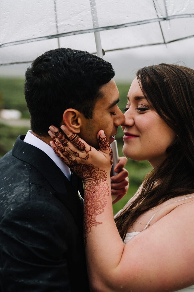 A bride leans in to kiss her groom. The bride has henna on her hand. The couple is standing under an umbrella and rain drops can be seen on the groom's black, wool suite. This couple had a family center elopement in the mountains of Idaho.