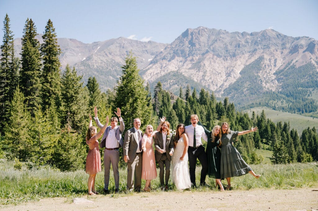 A bride and groom pose with their family after their Sun Valley, Idaho ceremony. The group has their hands in the air and they are cheering. There are mountains and trees behind the group.