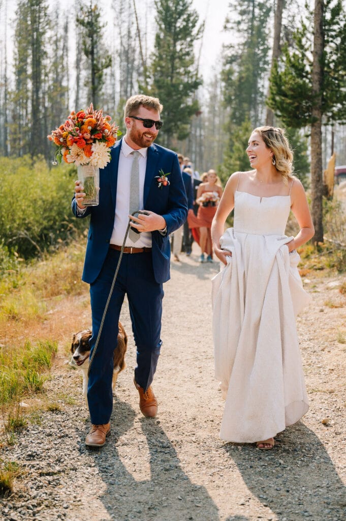 A bride and groom walk down a pathway toward their ceremony location. The couple is looking at each other and smiling. The groom is carrying a bouquet of flowers. The bride is holding up her dress. The couple's dog is walking beside them. The couple’s family is following in the background. This couple chose to elope with family in Stanley, Idaho.