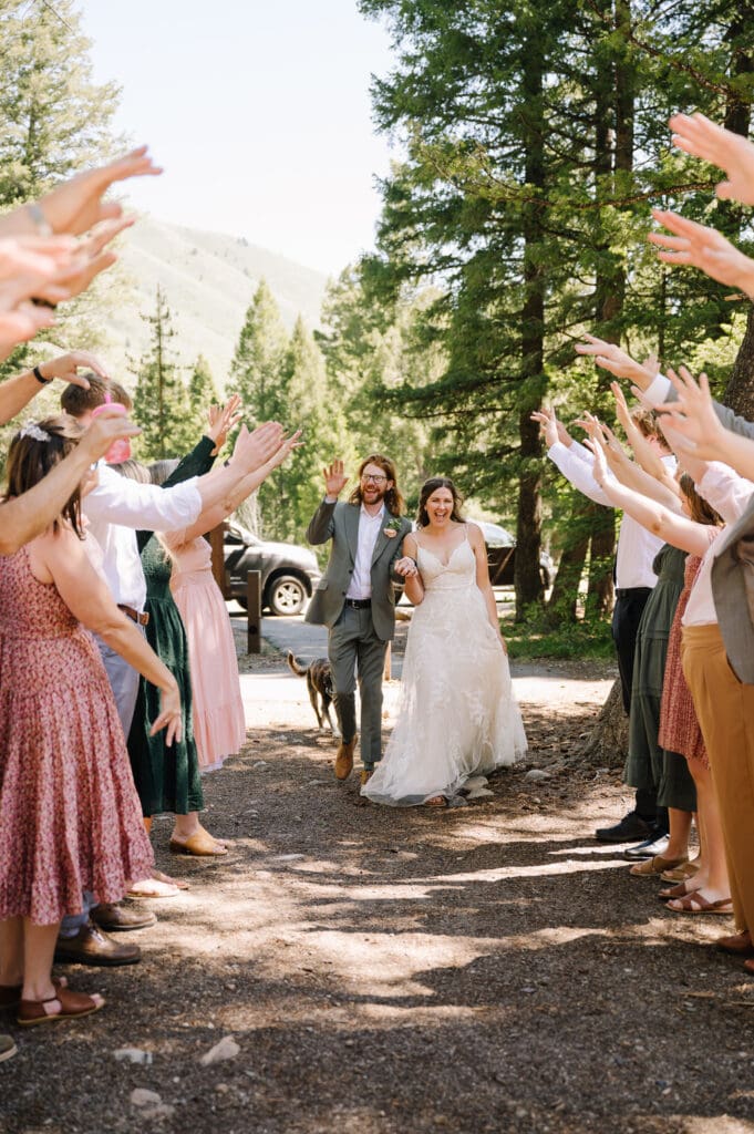 A couple walks through a greeting line at the entrance to their reception. The wedding guests are holding up their hands and the couple is walking underneath them. The couple is laughing with their guests. This couple chose to elope with family in a campground in Sun Valley.