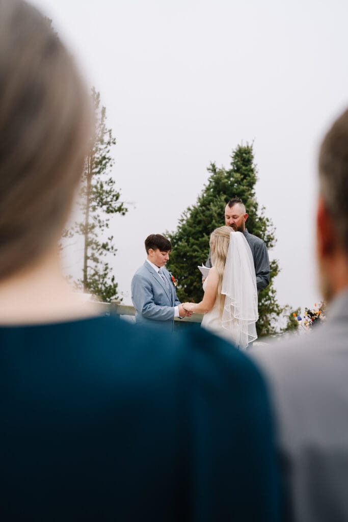 A couple stands holding hands during their sunrise ceremony with family. There are trees and fog behind the couple. This couple chose to have a sunrise ceremony with family and friends in Stanley, Idaho.