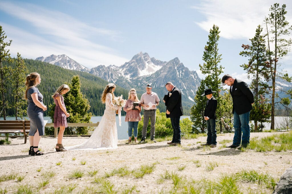 A couple and their wedding guests stand with their heads bowed in prayer. There are mountains and a lake behind them. This couple chose to elope with their family.