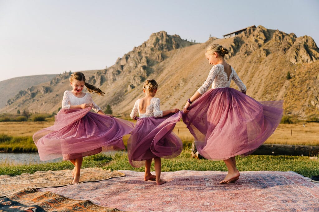 Three girls twirl in their flower girl dresses at an elopement reception. The dresses have white lace tops and blush colored tulle skirts. The girls are dancing on rugs and there are mountains behind them. 