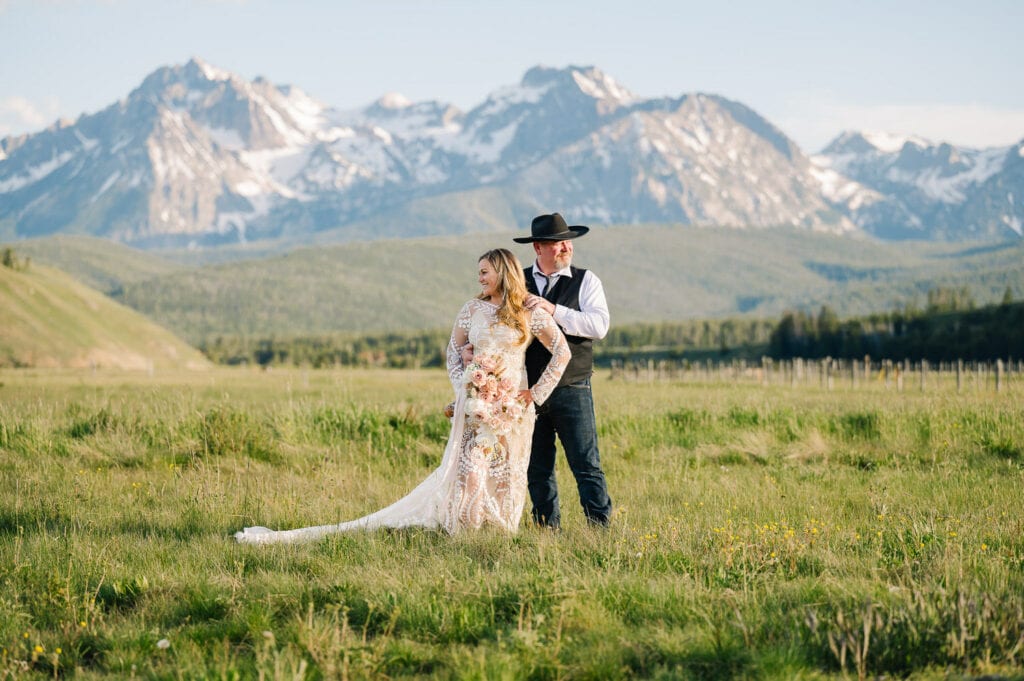 A couple stands in a green, grassy field with snow capped mountains rising behind them. The bride is wearing a long sleeved, lace wedding dress and holding a bouquet of flowers. The bouquet has white orchids and blush colored roses. The groom is wearing blue jeans, a black vest, and black cowboy hat. The couple is looking in opposite directions. This couple eloped with family in Stanley, Idaho.