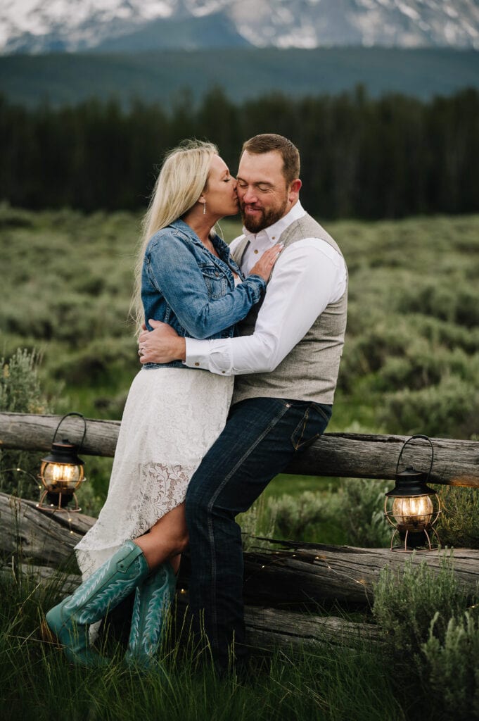 A groom leans against an old wood fence. His bride is leaning against him and kissing his cheek. The bride is wearing turquoise cowboy boots and a blue jean jacket over a white lace, knee-length wedding dress. The groom is wearing jeans, a white shirt, and gray vest. This couple eloped in the mountains of Idaho.