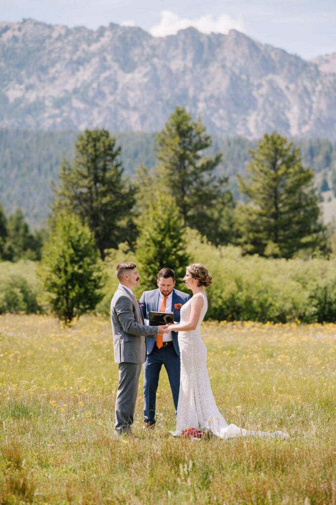 A couple stands with an officiant during the ceremony. The group is standing in a green, grassy field and there are mountains behind them. This couple chose to elope with a single wedding guest (their officiant). 