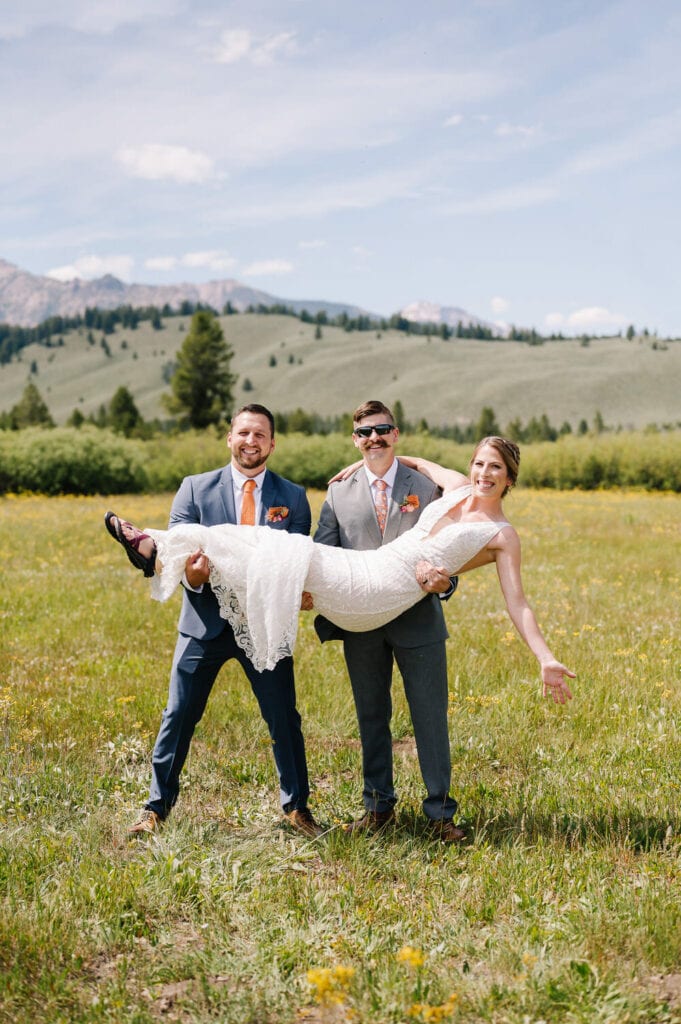 A couple smiles for the camera with their friend and Idaho officiant. The officiant and groom are holding the bride. The group is standing in a green, grassy field and there are mountains behind them.