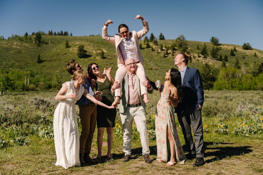 A bride and bride hang out with their wedding party. One bride is on the shoulders of a friend and the other bride is tickling her feet. The group is laughing together. One bride is wearing a pink suit with a bolo tie. The other bride is wearing a white dress.