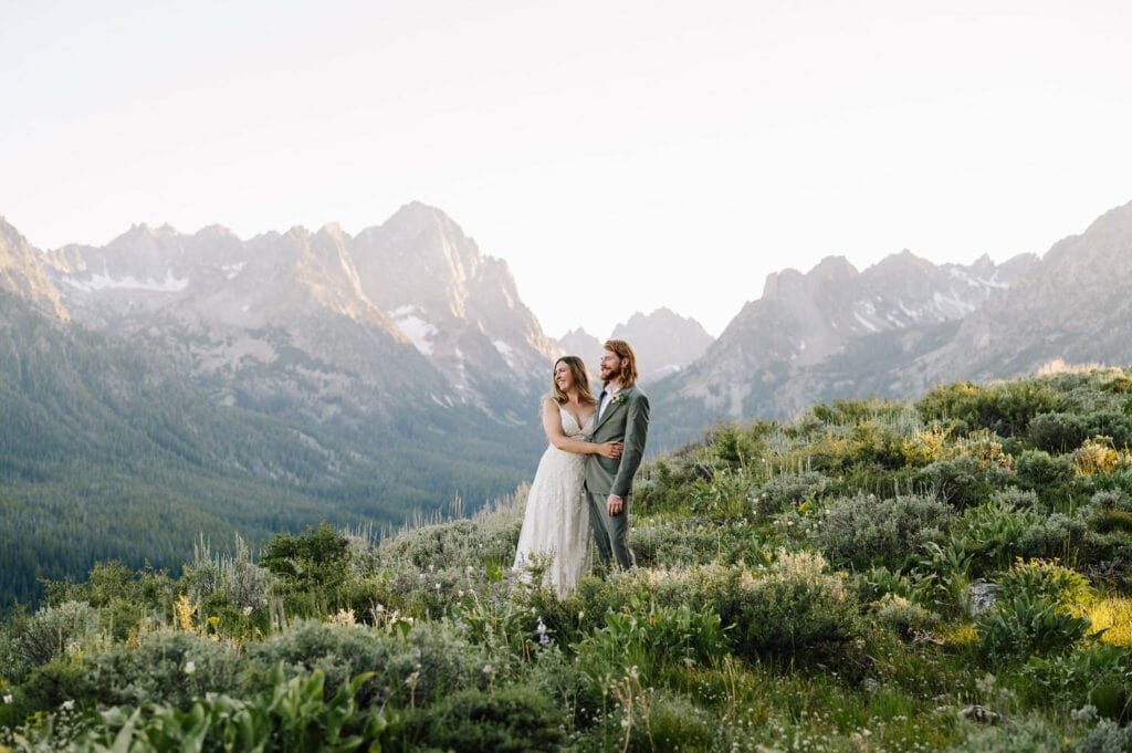 A bride and groom stand together on a ridge at sunset. There are mountains behind the couple. The room is wearing a sage green suit and the bride is wearing a white dress. This couple elope with family in Sun Valley and then drove to Stanley for an evening hike and private vow exchange in the mountains.