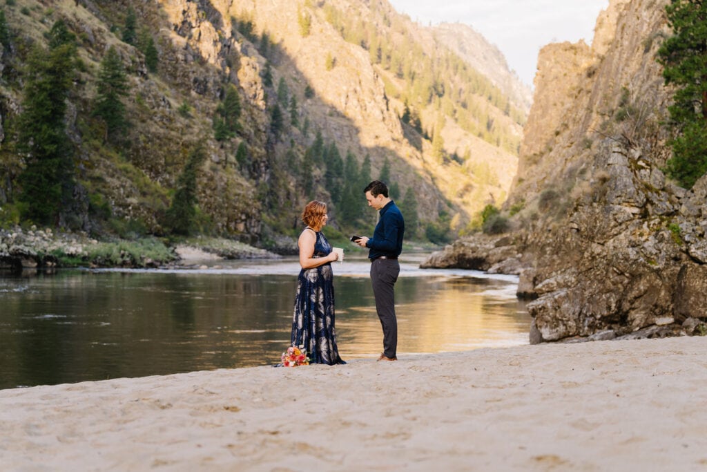 A couple exchanges vows in the early morning light. The groom is reading to his bride from a vow book. The couple is standing on a white sand beach with the Salmon River stretching behind them. She is wearing a blue dress with a silver flower pattern. He is wearing gray pants and a blue shirt.