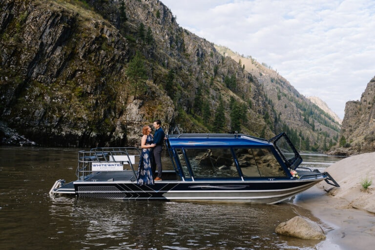 A bride and groom stand together on a jet boat on the Salmon River in Idaho. This couple eloped and took a private jet boat to their ceremony location. The boat is parked against a white sand beach.