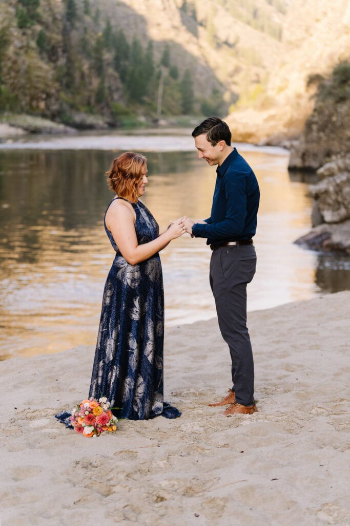 A couple exchanged rings during their elopement ceremony. The couple is standing on a beach and the Salmon River is behind them. 