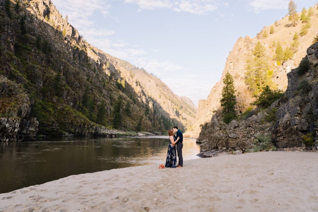 A bride and groom exchange their first kiss during their elopement ceremony. The sun is lighting up a large river and mountains behind the couple.