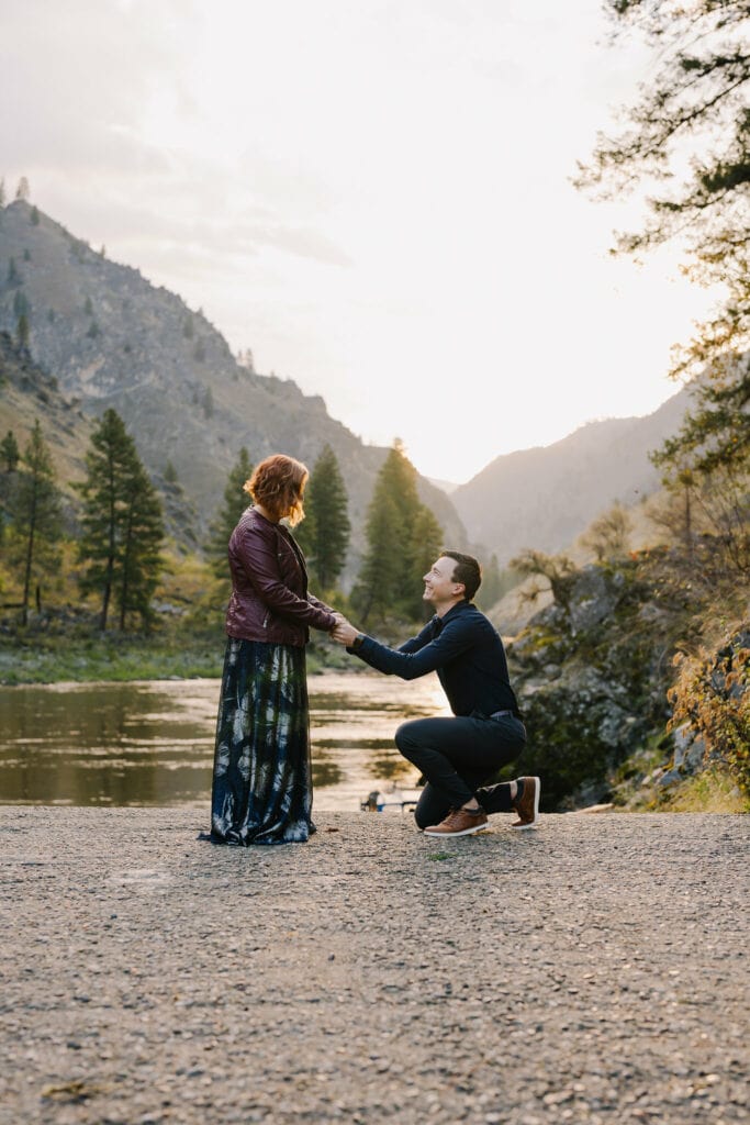 A groom proposes to his bride during their elopement. The couple is standing on a boat ramp with the Salmon River behind them. The groom is down on one knee.
