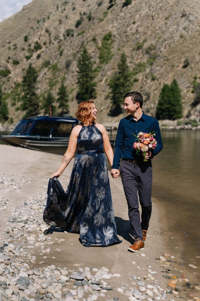 A bride and groom walk down a beach together on the Salmon River. The groom is holding an orange toned bouquet and the hand of his bride. The couple is laughing together. There is a blue jet boat in the background.