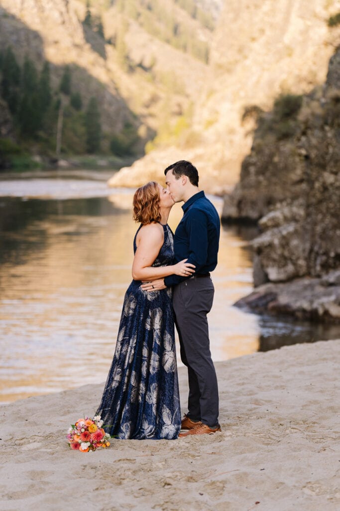 A bride and groom exchange their first kiss during the elopement ceremony on the banks of the Salmon River in Idaho. The couple is wearing blue toned outfits. An orange toned bouquet of fresh flowers lays at the bride's feet.
